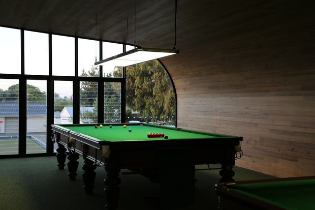 A dark, atmospheric billiards room with a full-size green pool table as the focal point. The room features extensive black-framed glazing, including a curved window, overlooking a suburban setting, plus a warm timber-lined wall and ceiling. A large suspended rectangular light illuminates the table, while a second pool table is partially visible in the foreground.