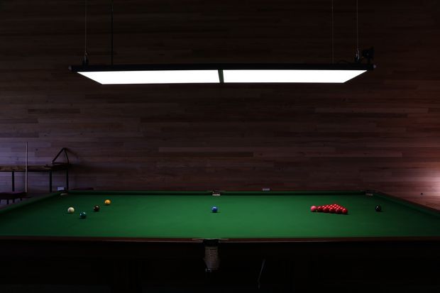 A dimly lit billiards room dominated by a full-size green-felt pool table, with billiard balls arranged on the table and a large rectangular pendant light above. The walls and ceiling are clad in dark horizontal timber boards, creating a moody, wood-lined recreational interior.