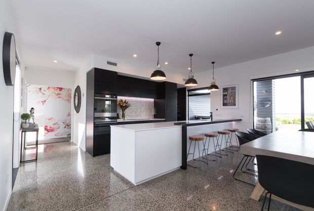 Bright contemporary open-plan kitchen and dining area with a large white island, black built-in cabinetry, integrated appliances, pendant lights, terrazzo flooring, and a dining table beside expansive windows. A floral artwork and small decorative furnishings add colour to the otherwise monochromatic space.