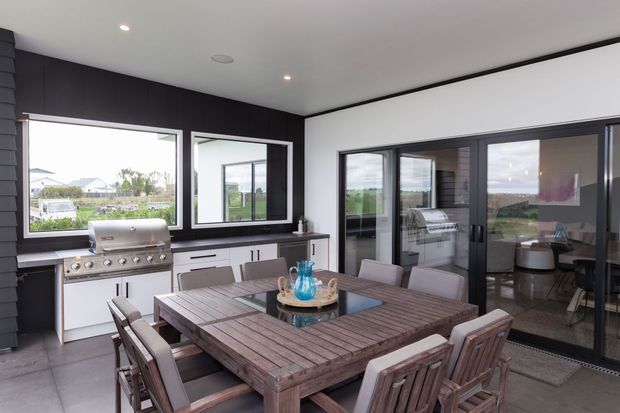 A contemporary enclosed outdoor entertaining area with a large timber dining table and chairs, built-in barbecue, dark cabinetry, expansive windows, and dark-framed sliding glass doors. The space opens visually toward a green suburban landscape and includes a glimpse of an adjacent interior living area through the glazing.