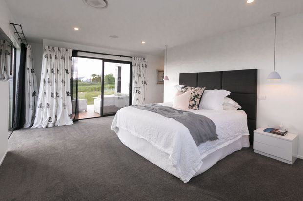 Bright contemporary master bedroom with a large upholstered dark headboard, layered white bedding, a grey throw, grey carpet, white walls, and minimalist white bedside furniture. Full-height sliding glass doors with patterned curtains open to an outdoor terrace, while pendant lights and recessed ceiling lights provide soft illumination.