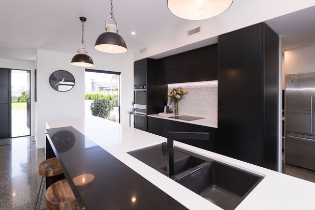 Contemporary kitchen with a large black-and-white island, integrated dark cabinetry, double undermount sink, stainless-steel appliances, and a pale textured feature splashback. Pendant lights hang above the island, with bar stools along one side and glazing opening toward an outdoor area.