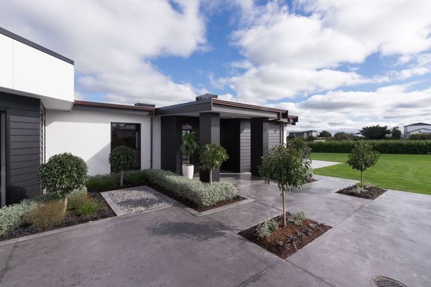 Contemporary single-storey residential exterior with low-profile flat roof forms, white rendered walls, dark horizontal cladding, and a broad concrete driveway or paved forecourt. Structured garden beds with clipped shrubs and young ornamental trees frame the entrance, with a large manicured lawn and hedge extending to the right.