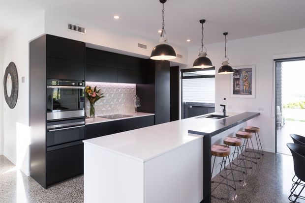 Contemporary kitchen with a large white island and integrated seating, matte black full-height cabinetry, built-in stainless-steel appliances, a pale geometric tiled splashback, and three black pendant lights. The kitchen opens toward a bright exterior through glazed doors, with terrazzo-style flooring and a small dining area visible at right.