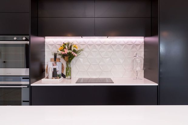 A sleek contemporary kitchen wall with matte dark cabinetry, integrated stainless-steel appliances, a white geometric textured tile splashback, pale benchtop, and a flush black induction cooktop. Soft pink under-cabinet lighting highlights the splashback, with a floral arrangement and glass cloche adding decorative detail.