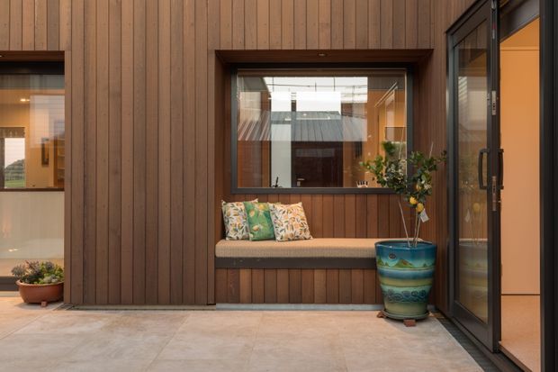 A warm, compact enclosed courtyard or entry space with vertical timber wall cladding, a built-in window seat beneath a large internal-facing window, and dark-framed glazed doors on the right. The seat has patterned cushions, with a decorative ceramic planter beside it. Pale large-format paving covers the floor, creating a sheltered, timber-lined outdoor living nook.