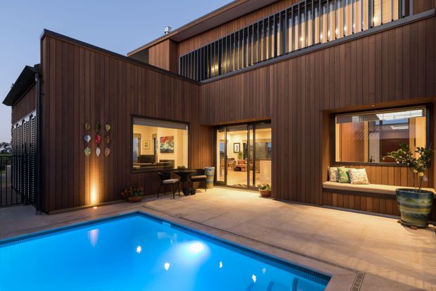 Exterior dusk view of a contemporary multi-level residence clad in dark vertical timber, arranged around a paved pool terrace. Large glazed openings reveal warmly lit interiors, while an upper-level screened balcony or clerestory zone adds strong horizontal and vertical architectural detailing. The illuminated swimming pool dominates the foreground.