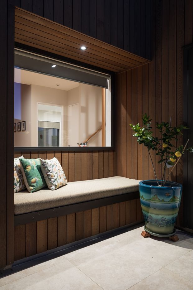 A compact enclosed courtyard or patio nook with dark vertical timber wall cladding, a built-in upholstered window seat, and a large decorative blue ceramic planter containing a citrus-like plant. A large opening with a roller blind looks into a bright interior hallway and stair area. The ceiling soffit is lined with warm timber boards and recessed downlights, while the floor is finished in large pale tiles.