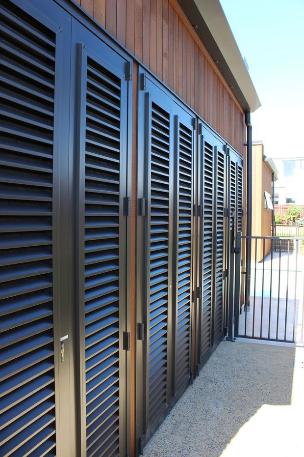 Exterior view of a contemporary building façade with a long row of dark louvred shutter doors or screens set against vertical timber cladding. A narrow concrete or terrazzo-finished walkway runs alongside the façade, with a simple dark metal gate and railing at the end. The image emphasizes ventilation shutters, robust exterior joinery, and clean linear detailing in bright daylight.