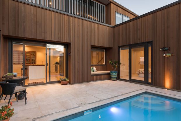 A contemporary multi-level courtyard exterior clad in vertical dark timber, with large glazed sliding doors opening to warm interior spaces. The paved terrace surrounds a swimming pool, with outdoor dining furniture, potted plants, integrated lighting, and a small upper-level balcony with vertical metal balustrades.