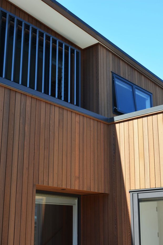 Close-up exterior view of a contemporary multi-level building clad in vertical timber boards. The facade features dark-framed windows, a projecting upper-level balcony or screen with evenly spaced vertical metal slats, and clean roof and wall junctions. Strong sunlight highlights the warm wood tones against a clear blue sky.