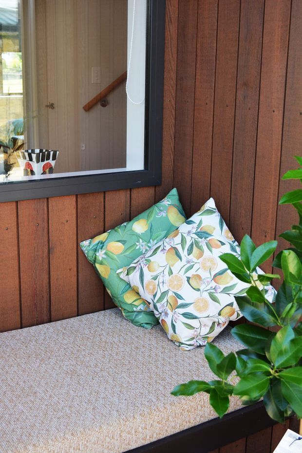 A compact built-in window seat or bench is shown against a vertical dark timber-paneled wall. The seat has a textured beige upholstered cushion and two colourful botanical cushions with citrus and leafy motifs. A black-framed window reflects an interior stair and timber handrail, while a leafy plant occupies the foreground. The scene has a warm, relaxed, nature-inspired residential feel.