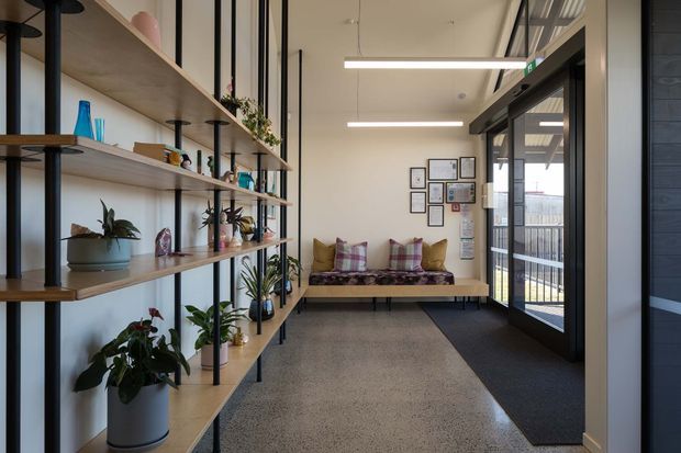 A bright, contemporary entry or reception area with open timber display shelving supported by slim black metal posts along the left wall. The shelves hold plants, books, pottery and decorative objects. A built-in bench with patterned cushions sits beneath framed artwork at the far end, while a glazed dark-framed doorway opens to an exterior balcony or walkway. Linear pendant lighting, polished concrete or terrazzo-style flooring, and white walls create a clean, welcoming atmosphere.