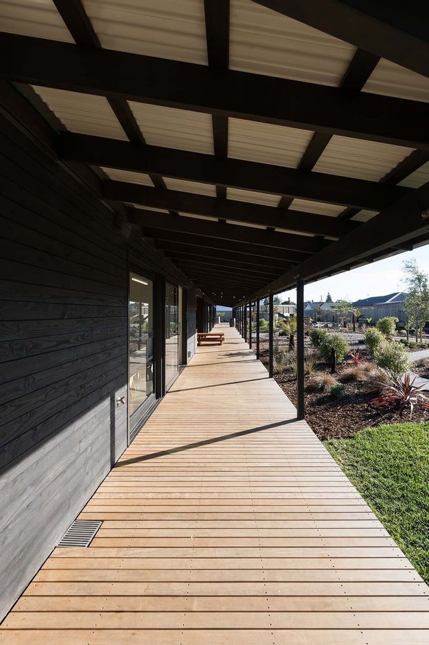 A long covered exterior walkway runs alongside a dark-clad building. The path has a timber deck, black structural posts and beams, and a translucent corrugated roof supported by exposed rafters. Glazed doors open from the building onto the deck, while landscaped garden beds and neighbouring low-rise buildings are visible to the side.