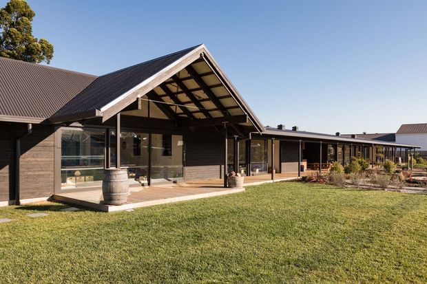 Single-storey rural-style building viewed from a broad lawn, featuring dark horizontal cladding, a prominent pitched gable roof with exposed timber rafters, a covered timber deck, and extensive glazing. The long low wing extends to the right, with simple landscaping and barrel planters near the veranda.