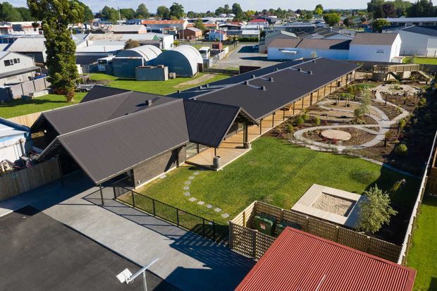 Aerial view of a low-rise institutional or commercial complex with multiple dark metal pitched roofs, covered walkways, landscaped courtyards, curved garden paths, lawns, and surrounding suburban buildings. The site includes several curved-roof structures and a fenced service or recreational area.