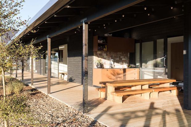 Single-storey contemporary home with a deep covered outdoor veranda, timber decking, dark timber cladding and exposed roof structure. The veranda includes a built-in outdoor kitchen or bar with a stone-and-brick-look splashback, timber cabinetry, and a substantial timber dining table with bench seating. Glazed sliding doors connect the covered terrace to the interior, while landscaped planting and gravel border the deck.