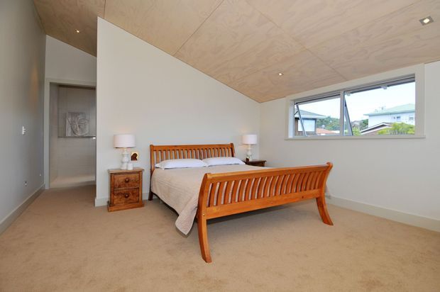 A bright, simply furnished bedroom with a traditional timber bed featuring slatted headboard and footboard, matching timber bedside tables, beige carpet, white walls, and a pale timber-lined ceiling. A large horizontal window provides views to neighbouring homes, while a doorway opens to an adjoining area.