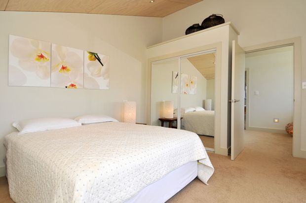 Bright, calm bedroom with a large bed dressed in white bedding, pale neutral walls, beige carpet, and a light timber ceiling. A mirrored built-in wardrobe reflects the bed and floral wall artwork, while an open doorway leads to a neutral hallway or adjacent room. Soft bedside lighting and minimal decor create an airy, serene feel.