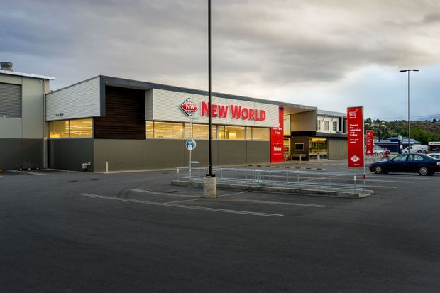 Exterior view of a New World supermarket in a paved car park. The building has a contemporary low-rise commercial facade with grey horizontal metal cladding, dark recessed timber-look wall panels, extensive storefront glazing, prominent red New World signage, and covered entry canopies. Parking bays, accessibility railings, signage, and a few vehicles are visible in the foreground and background.