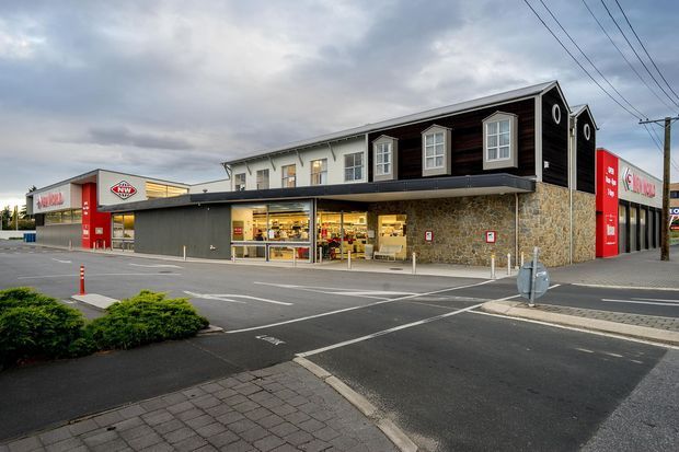 Exterior street-level view of a contemporary commercial building, likely a retail or convenience-store development. The building combines dark horizontal cladding and white upper walls with stone veneer, large glazed shopfronts, red branded signage, and a broad paved parking and pedestrian forecourt. Overhead utility lines and a cloudy sky are visible.