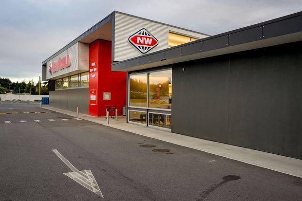 Exterior view of a modern New World supermarket building, featuring dark cladding, a bright red entrance tower, horizontal metal facade panels, extensive glazing, and prominent New World signage. The foreground shows a paved vehicle access area with directional markings.