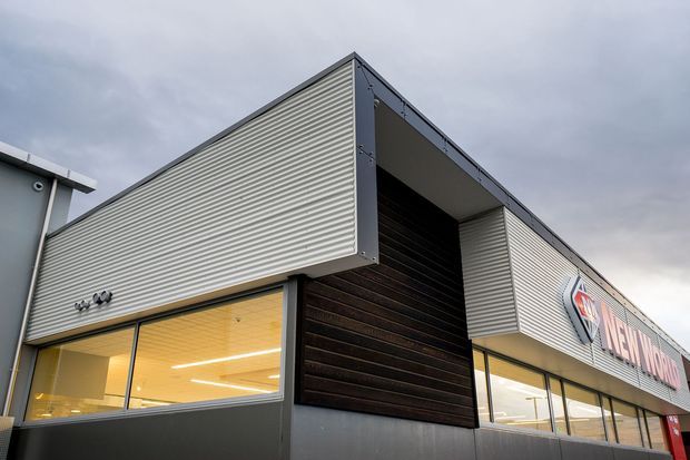 Exterior view of a contemporary commercial building with a crisp angular form, corrugated light-grey metal cladding, dark vertical feature cladding, charcoal framing, and large illuminated glazed windows. A prominent retail or business sign is visible along the facade beneath an overcast sky.
