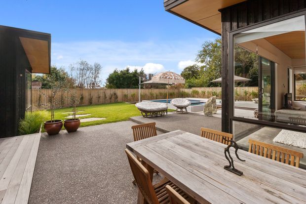 A contemporary outdoor dining terrace overlooking a landscaped backyard and swimming pool. The space features a large weathered timber dining table with wooden chairs, dark timber-clad building elements, broad glazed openings, a covered eave, paved surfaces, lawn, outdoor lounge seating and a pool beyond.