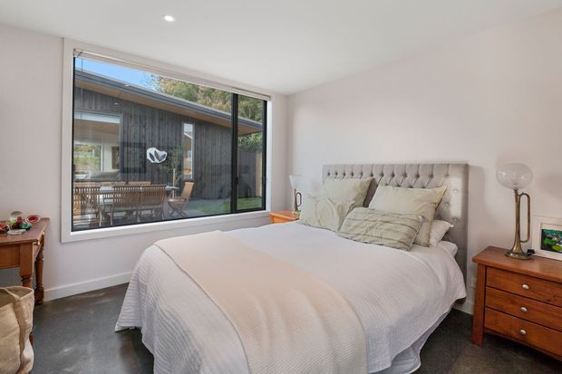 Bright, contemporary bedroom with a tufted upholstered bed, layered neutral bedding, timber bedside tables, and a large picture window overlooking a covered outdoor courtyard or deck. The room has white walls, recessed ceiling lighting, and a dark smooth floor.