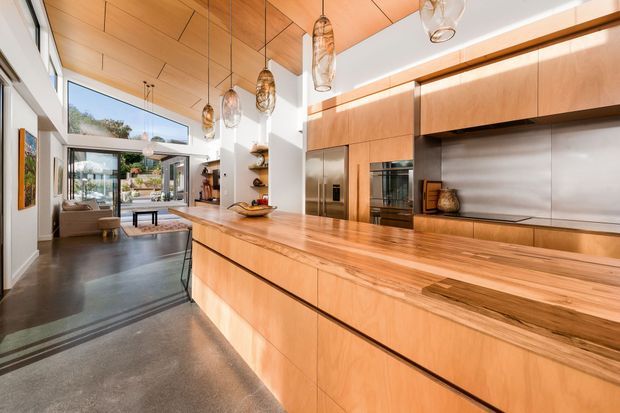 A spacious open-plan kitchen with extensive light timber cabinetry and a large timber island. The kitchen features a stainless-steel splashback, integrated stainless appliances, polished concrete flooring, a timber-lined ceiling, and multiple sculptural pendant lights. Full-height glazing and open doors connect the kitchen to a bright living area and exterior.