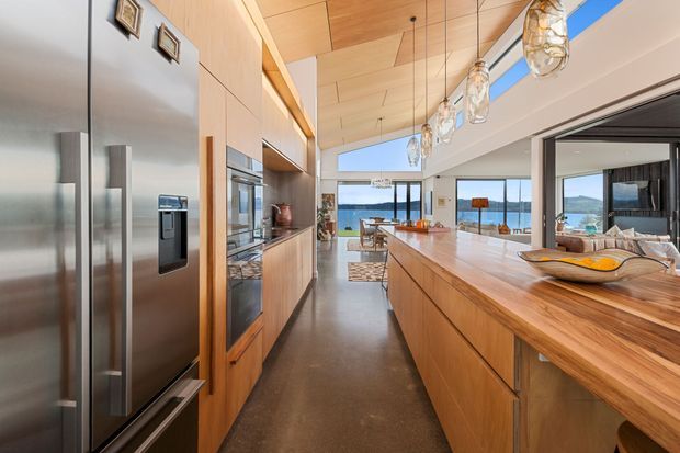 A spacious contemporary kitchen with extensive warm timber cabinetry and a large timber island, opening toward a bright living area and expansive waterfront glazing. Stainless steel appliances, pendant lights, polished concrete flooring, and a linear timber ceiling create a refined, open-plan interior.