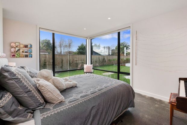 A bright contemporary bedroom with a large bed dressed in layered grey and white bedding, floor-to-ceiling corner glazing, and views onto a small fenced lawn. A compact piano and wall-mounted display shelving add personal character, while recessed ceiling lights and a minimalist white interior keep the space clean and airy.