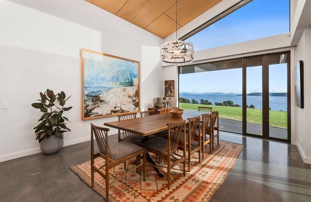 Bright dining room with a long timber dining table and matching spindle-back chairs arranged on a patterned rug. The room features polished concrete flooring, white walls, a warm timber-lined sloped ceiling, pendant lighting, and expansive dark-framed sliding glass doors opening to a lawn and expansive water view.