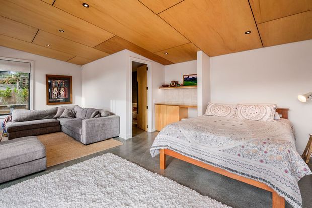 A bright open-plan bedroom and living area with a timber-paneled ceiling, white walls, grey sectional sofa, upholstered bed, pale patterned bedding, and a built-in timber desk or shelving unit. Large windows bring in natural light, while recessed ceiling lighting and warm wood finishes create a comfortable contemporary interior.