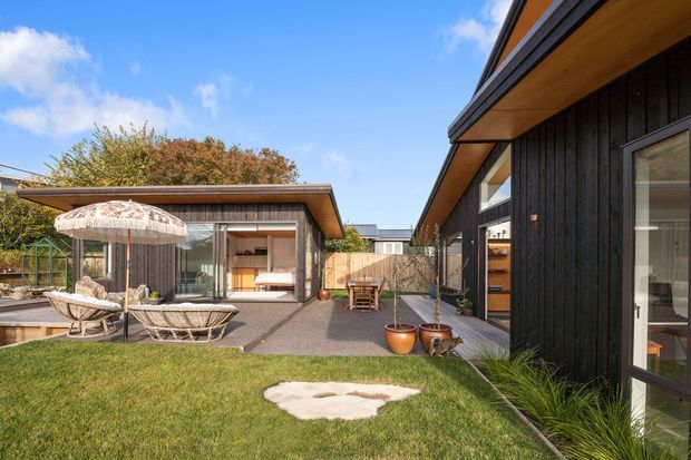 A contemporary residential courtyard viewed from the garden, with two dark timber-clad low-rise structures framing a lawn and paved outdoor area. The buildings have low-pitched rooflines, broad timber soffits, large glazed openings and timber-framed doors. Outdoor lounge furniture and potted plants create a relaxed, modern garden setting.