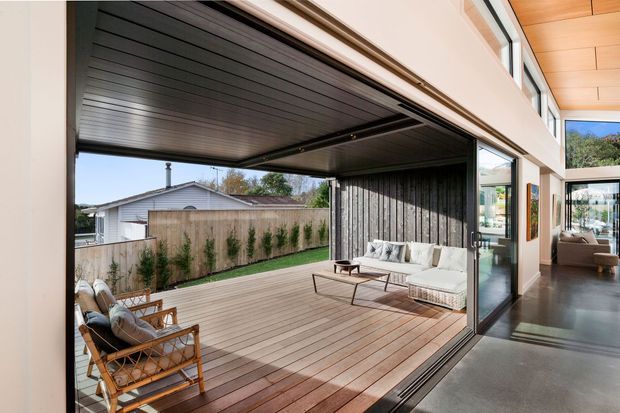 Contemporary indoor-outdoor living space with a large retractable glazed opening connecting a polished interior to a timber-decked outdoor lounge. The covered terrace features lounge seating, a coffee table, timber decking, and a dark slatted ceiling, with a fenced garden and neighbouring house beyond. The interior has expansive glazing, dark flooring, and a warm timber ceiling.
