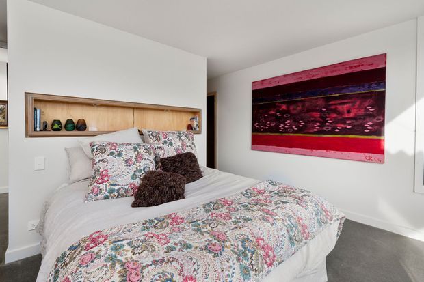 Bright bedroom with a large bed dressed in a floral patterned quilt and matching pillows, a recessed timber display niche in the wall, white walls, grey carpet, and a bold abstract artwork above the bed.