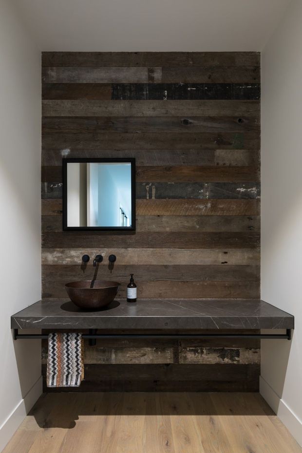 A compact bathroom vanity area featuring a wall-mounted dark reclaimed timber feature wall, a dark stone benchtop, a round vessel basin, black wall-mounted tapware, a framed mirror, and an open black metal vanity frame with a hanging patterned towel. Light timber flooring and white side walls provide contrast.