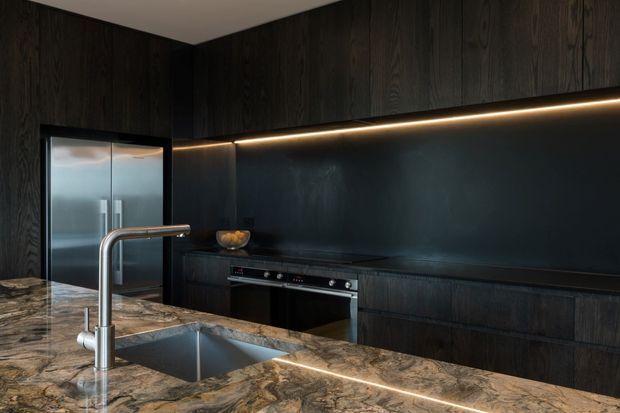A dark, sophisticated contemporary kitchen featuring extensive black-stained timber cabinetry, a matching dark splashback, integrated appliances, and a large island with a dramatic natural stone benchtop. A stainless-steel sink and gooseneck tap sit in the foreground, with linear under-cabinet lighting creating a warm accent.
