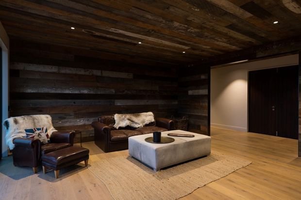 A dark, rustic living room with reclaimed timber wall and ceiling panelling, leather lounge chairs and sofa, a large upholstered ottoman, and a natural woven rug. Warm timber flooring and recessed ceiling lighting create a cozy, moody atmosphere, with a simple hallway and dark entry door visible to the right.