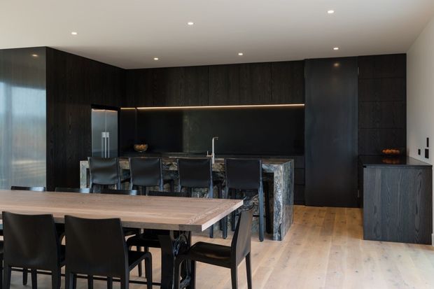 A sophisticated open-plan kitchen and dining area with dark timber cabinetry, a black stone island and splashback, integrated appliances, and warm light timber flooring. A large timber dining table with black upholstered chairs occupies the foreground, while recessed ceiling lights and concealed shelf lighting create a refined, moody atmosphere.