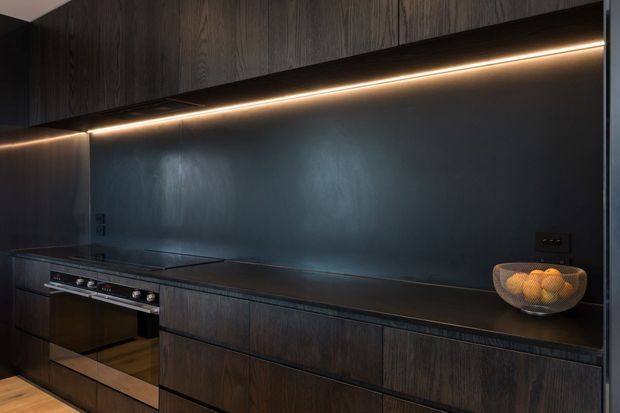 A dark, contemporary galley-style kitchen with full-height charcoal timber cabinetry, a long black benchtop, integrated cooktop, built-in oven, and a dark reflective splashback. Warm linear under-cabinet lighting highlights the joinery and benchtop, creating a moody, refined atmosphere.
