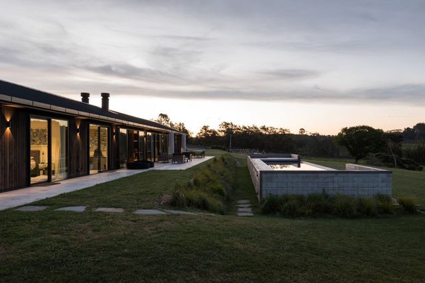 A low, elongated modern residence set in a landscaped rural or semi-rural setting at dusk. The dark-clad building has extensive floor-to-ceiling glazing, a low-pitched roof, and an outdoor terrace with seating. A rectangular concrete-block swimming pool structure occupies the foreground, surrounded by lawn and native planting.