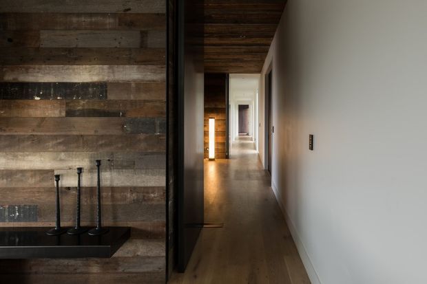A long, narrow residential hallway with warm timber flooring and a dark exposed timber ceiling. The right wall is plain white with a small black switch plate, while the left side features a dark timber feature wall and a black built-in fireplace or display ledge with slender vertical elements. Recessed doorways and warm lighting lead toward a brighter room at the end, creating a moody, understated interior.