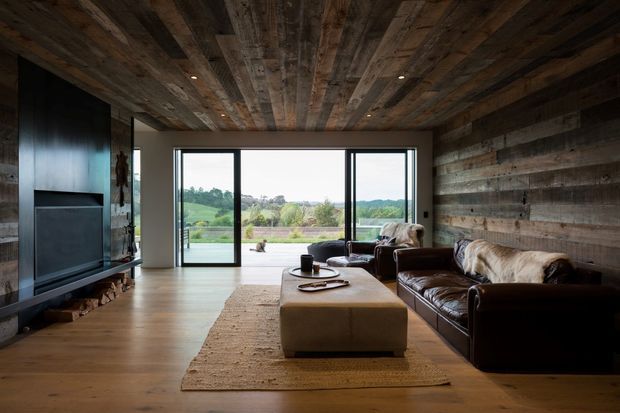 Rustic contemporary living room with reclaimed timber walls and ceiling, warm timber flooring, a large dark built-in media unit, leather sofas, and a central upholstered coffee table. Full-height sliding glass doors open to a terrace and expansive green rural landscape beyond.