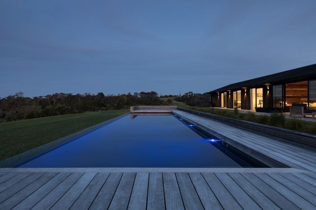 Twilight exterior view of a long, linear modern residence beside a narrow illuminated swimming pool. A broad timber deck surrounds the pool, while the low-slung dark-clad house has extensive warm-lit glazing and a projecting roofline. Open lawn and tree-covered landscape extend beyond the property.