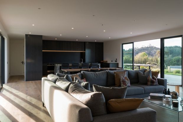 A spacious contemporary open-plan living room with a large sectional sofa in the foreground, a dark built-in kitchen or bar joinery wall behind it, and expansive floor-to-ceiling glazing overlooking a green landscape. Recessed ceiling lights, warm timber-toned flooring, dark cabinetry, and abundant natural light create a refined, calm interior.