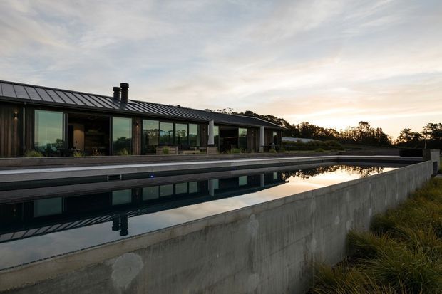 Low, elongated contemporary house with a dark metal pitched roof, extensive floor-to-ceiling glazing, and a long reflecting or swimming pool in the foreground. The concrete pool wall and natural grasses frame the house, which is set against a wooded landscape in soft evening light.