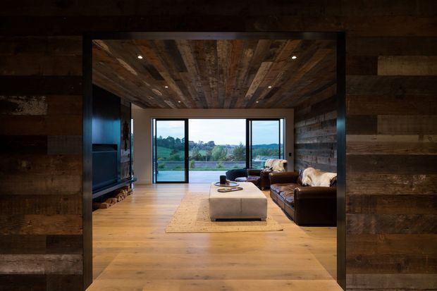 Warm, rustic living room framed by a dark timber opening, featuring extensive reclaimed timber wall and ceiling cladding, wide timber flooring, a large glazed opening to the landscape, a dark leather sofa, and a central upholstered ottoman on a neutral rug. The room has a relaxed lodge-like atmosphere with integrated ceiling downlights and strong indoor-outdoor connection.