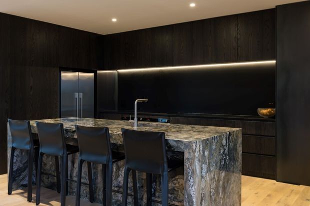 A dramatic contemporary kitchen with dark vertical timber cabinetry, a large waterfall island clad in heavily veined natural stone, integrated appliances, a black splashback, and four black bar stools. Warm recessed and linear lighting contrasts with the dark finishes, while pale timber flooring adds brightness.